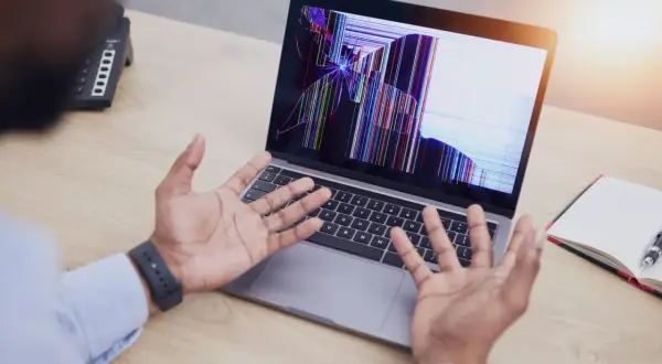 Rozlany ekran w laptopie wyświetla kolorowe pęknięcia, a osoba z rozłożonymi rękami patrzy na niego z niedowierzaniem.