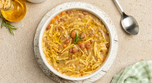 Un bol de pasta e fagioli, une soupe réconfortante avec des pâtes fines et des haricots, garnie de romarin. Une cuillère et une petite bouteille d'huile d'olive sont à proximité.