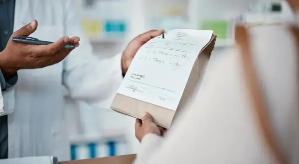 Farmaceuta w białym fartuchu podaje pacjentowi receptę i torebkę z lekami.