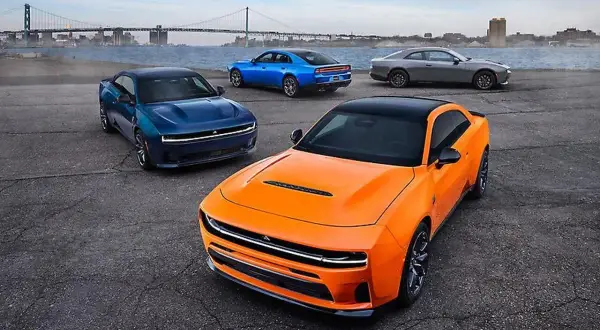 Pomarańczowy i niebieski Dodge Charger, szary Dodge Challenger i niebieski Dodge Charger stoją nad wodą z mostem w tle.