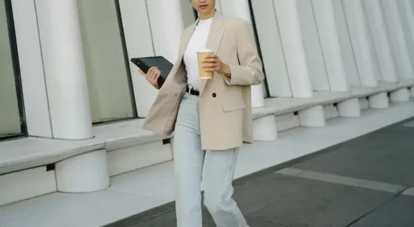 Eine Frau im smart casual Look mit beigem Blazer, Jeans und Kaffee in der Hand, geht mit einem Tablet.