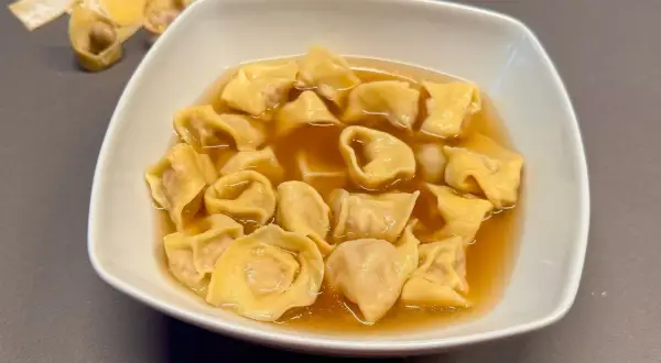 Un piatto fumante di tortellini in brodo, un classico confortante. Sullo sfondo, alcuni tortellini crudi e sfoglia.
