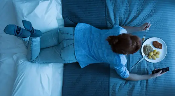 A woman eats dinner in bed, holding a remote. Wondering how long should you wait to sleep after eating?