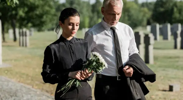Para idąca przez cmentarz, kobieta w czerni z bukietem kwiatów, mężczyzna w białej koszuli z marynarką.