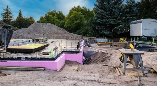Budowa w stanie zerowym: fundamenty z różową izolacją, zbrojenie, sterta piasku i sprzęt budowlany.