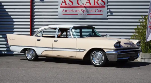 En krämvit och ljusbeige De Soto från 1950-talet står parkerad framför en byggnad med skylten "AS CARS".
