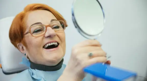 Uśmiechnięta kobieta z rudymi włosami ogląda swoje nowe zęby w lusterku. NFZ dofinansowanie do protez zębowych pozwoliło jej odzyskać pewność siebie.