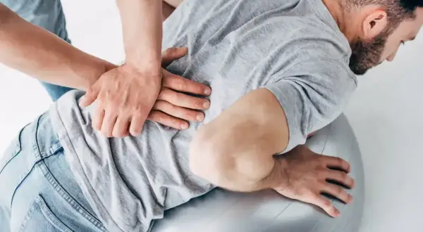Terapeuta wykonuje masaż pleców mężczyzny na piłce rehabilitacyjnej. Czy osteopata jest lekarzem? To pytanie nurtuje wielu pacjentów.
