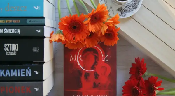 Stos książek, pomarańczowe gerbery i kubek kawy. Na okładce jednej z nich widnieje "Czarna Madonna" i tytuł "Mróz".