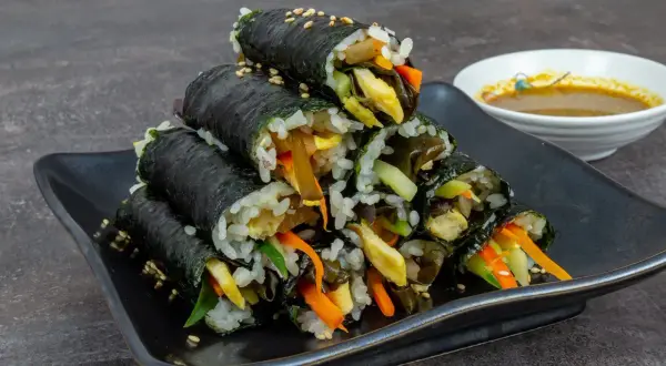 Gestapeltes Kimbap, koreanische Reisrollen mit Gemüse und Ei, auf schwarzer Platte, daneben eine Schale Dip.