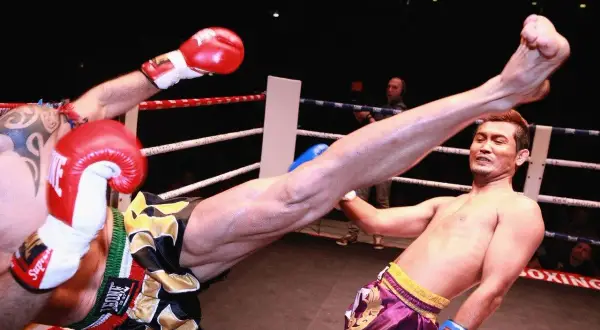 A fighter's leg swings high in a Muay Thai match. The opponent's flinching reflex is evident as he attempts to block the powerful kick.