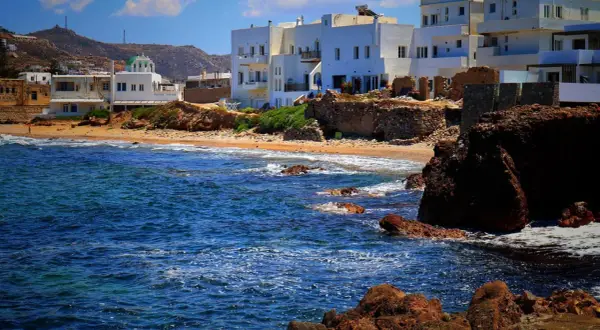 Blågrönt hav sköljer mot klippor och sandstrand. Vita hus med gröna tak reser sig mot en bergig bakgrund på Naxos, Grekland.