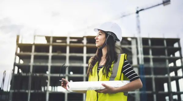 Junge Frau mit Helm und Warnweste auf Baustelle, hält Baupläne. Sie verfolgt ihren Weg im Bauingenieurwesen berufsbegleitend.
