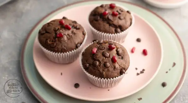 Drei saftige, vegane Muffins mit Schokostückchen und Granatapfelkernen auf einem rosa Teller.