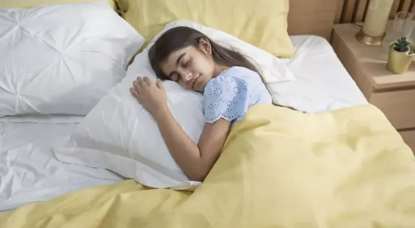 A young girl sleeps peacefully under a yellow blanket. This serene scene reminds us of the importance of fresh air and how to get rid of off-gassing smells for a healthy sleep environment.