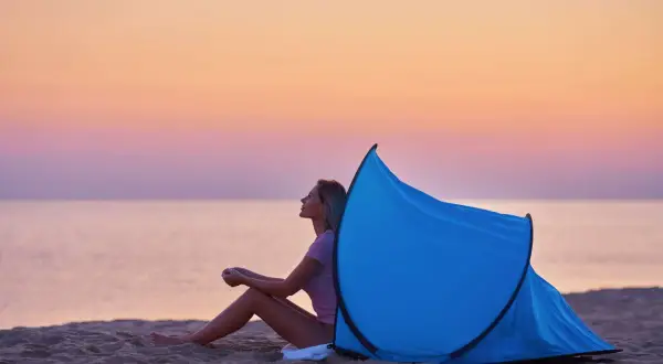 Kobieta siedzi na plaży przy rozłożonym namiocie plażowym. Zachód słońca nad morzem. Jak złożyć namiot plażowy?