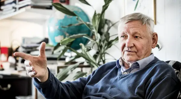 Äldre man med grått hår, liknar Ragnar Skanåker, gestikulerar med handen. Bakom honom syns en jordglob och växter.