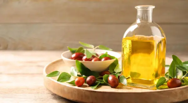 Flasche mit goldenem Simmondsia Chinensis Seed Oil neben einer Schale mit Jojobasamen und grünen Blättern auf einem Holzteller.
