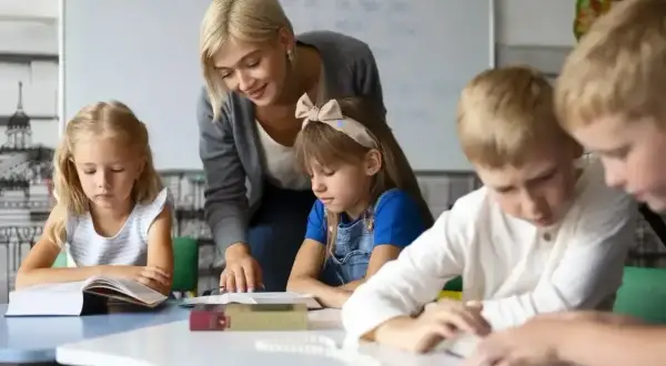 Nauczycielka wspiera uczniów w nauce, pokazując swoje kompetencje rady pedagogicznej. Dzieci skupione na książkach.