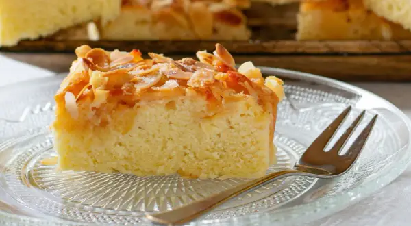 Ein Stück saftiger **Mandelkuchen vom Blech** mit knusprigen Mandelblättchen und Zucker bestreut, auf einem Glas-Teller mit einer Gabel.