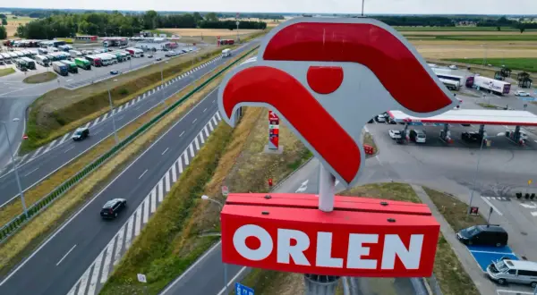 Duży znak ORLEN nad stacją paliw przy autostradzie. W tle parking z ciężarówkami i samochodami.