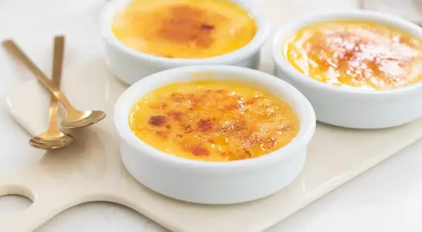 Tre krämiga creme brulee i vita skålar med karamelliserat sockerlock. Guldfärgade skedar ligger bredvid.