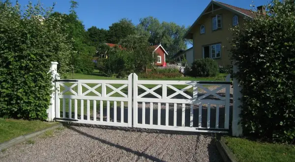 Vit grind med korsmönster leder in på en grusgång entre till ett gult hus och en röd stuga.