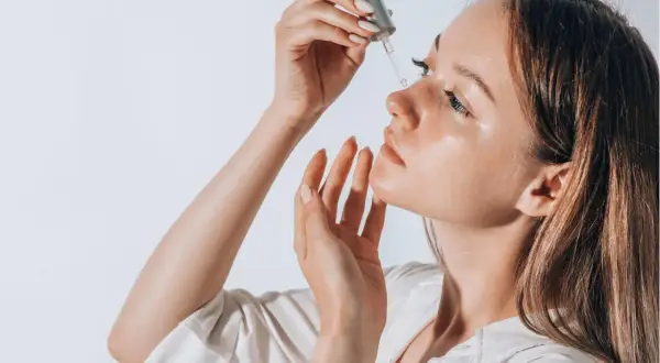 Frauenhand trägt Tropfen eines Serums auf das Gesicht auf. Ob Squalan oder Hyaluron, die Haut strahlt Feuchtigkeit aus.
