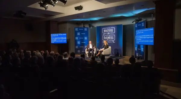 Conference photography captures a panel discussion on a stage with two speakers, audience members in the foreground, and Bloomberg branding.