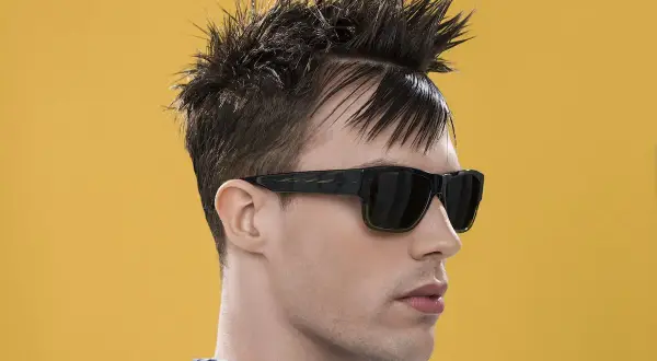 Uomo con capelli a spazzola corti, occhiali da sole scuri e camicia a quadri blu e bianchi su sfondo giallo.