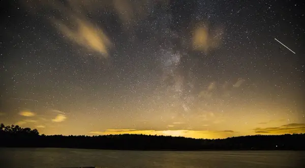 Nocne niebo nad jeziorem, pełne gwiazd i smug chmur, idealne do nauki, jak fotografować gwiazdy. Widać Drogę Mleczną i spadające gwiazdy.