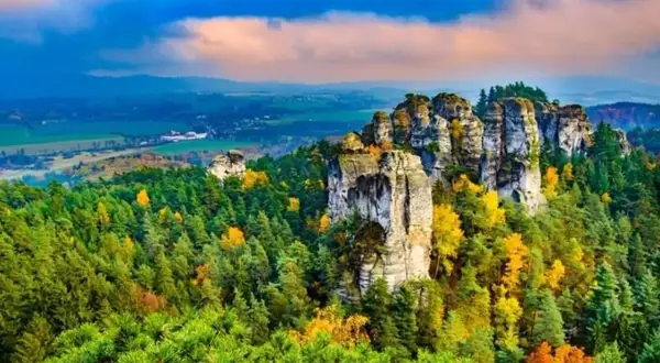 Felsformationen im böhmischen Paradies (Český ráj) ragen aus einem bunten Herbstwald hervor, unter einem dramatischen Himmel.