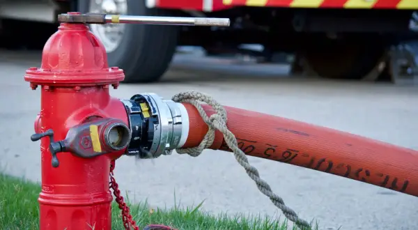 Czerwony hydrant naziemny podłączony do węża strażackiego, z tyłu widać fragment wozu strażackiego.