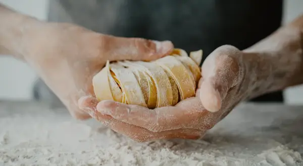 Dłonie trzymające świeży, pokrojony makaron tagliatelle, oprószony mąką.