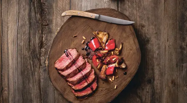 Saftige Tagliata di Manzo mit gebratenen Pilzen und Paprika auf einem rustikalen Holzbrett, garniert mit Pinienkernen und Balsamico.