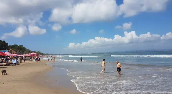 Słoneczny dzień na plaży w Seminyak na Bali. Ludzie spacerują po piasku i kąpią się w oceanie.