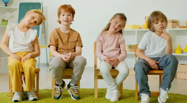 Cztery dzieci siedzą na krzesłach. Jedno z nich, z rudymi włosami, uśmiecha się, być może zmagając się z zaburzeniami neurorozwojowymi.