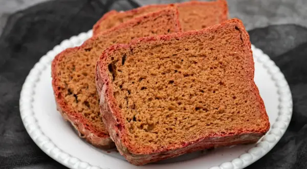 Kilka kromek pysznego, ciemnego chleba na zakwasie z chrupiącą, czerwonawą skórką, ułożonych na białym talerzu.