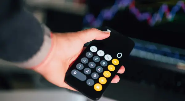 Dłoń trzyma smartfon z kalkulatorem, na ekranie widać wykresy finansowe. Obliczanie faktury bez vat.