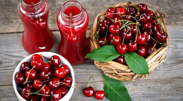 Dwa szklane dzbanki z rubinowym sokiem z czereśni, obok koszyk pełen świeżych owoców i miseczka z czereśniami.
