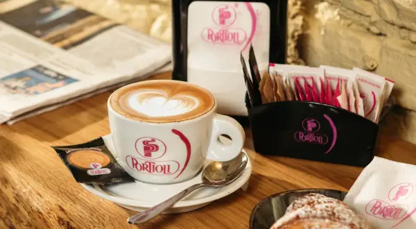 Un cappuccino senza schiuma, con un cuore di latte art, accompagnato da un croissant e un giornale.