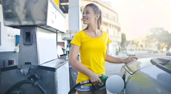 Uśmiechnięta dziewczyna tankuje auto, a w tle widać stację benzynową. Może to Quebonafide & Omega Sapien sniffing gas?