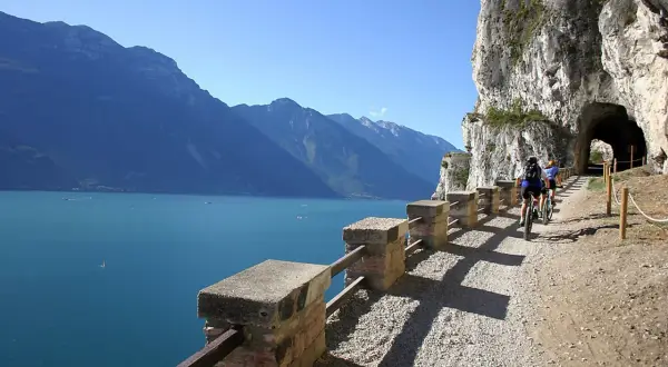 Ciclisti sul sentiero della Ponale, con il lago e le montagne sullo sfondo. Un tunnel scavato nella roccia attende.