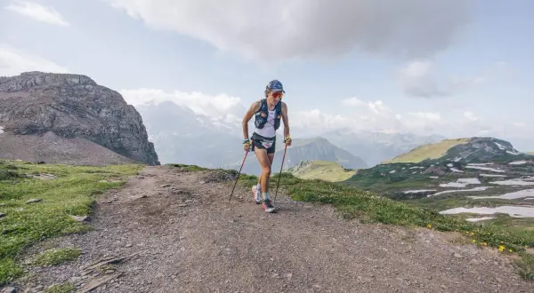 Corredor/a de montaña en un sendero rocoso, con bastones y vistas alpinas. Esto es trail running, pura aventura.