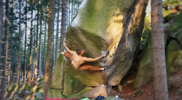 Mężczyzna wykonuje dynamiczny ruch podczas treningu wspinaczkowego w domu, na naturalnej skale w lesie.