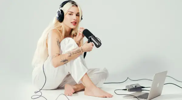 Elena Rose, a blonde woman with tattoos, sits on the floor wearing headphones and holding a microphone, ready to record.