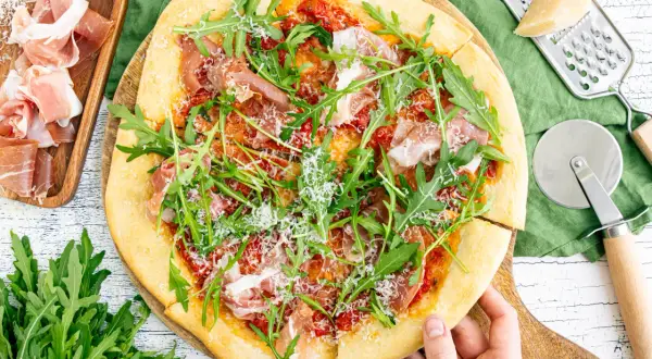 Pizza mit Rucola, Schinken und Parmesan, daneben Prosciutto, Parmesan und Pizzaschneider.
