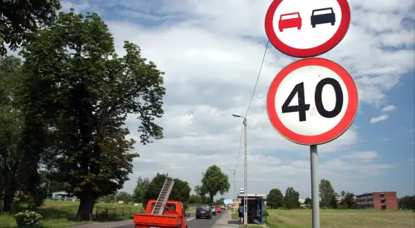 Znak zakazu wyprzedzania i ograniczenie prędkości do 40 km/h. Pomarańczowy pick-up z drabiną jedzie po drodze.