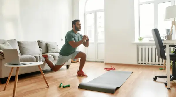 Un homme fait des fentes dans son salon, une idée pour comment commencer le sport à la maison. Un tapis, des haltères et un ordinateur sont à proximité.