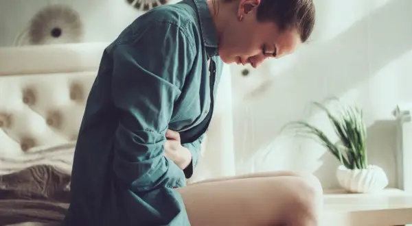 Kobieta w niebieskiej koszuli trzyma się za brzuch, cierpiąc z powodu bólu. Zastanawia się, ile trwa okres i jak sobie z nim poradzić.